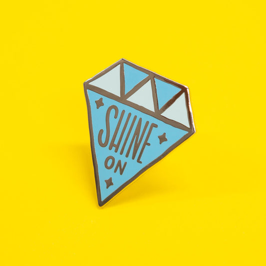 diamond shaped enamel pin that says "shine on"